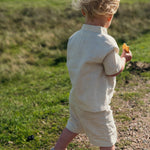 Back view of a baby wearing a beige short linen shirt and matching shorts while walking on a grassy path.