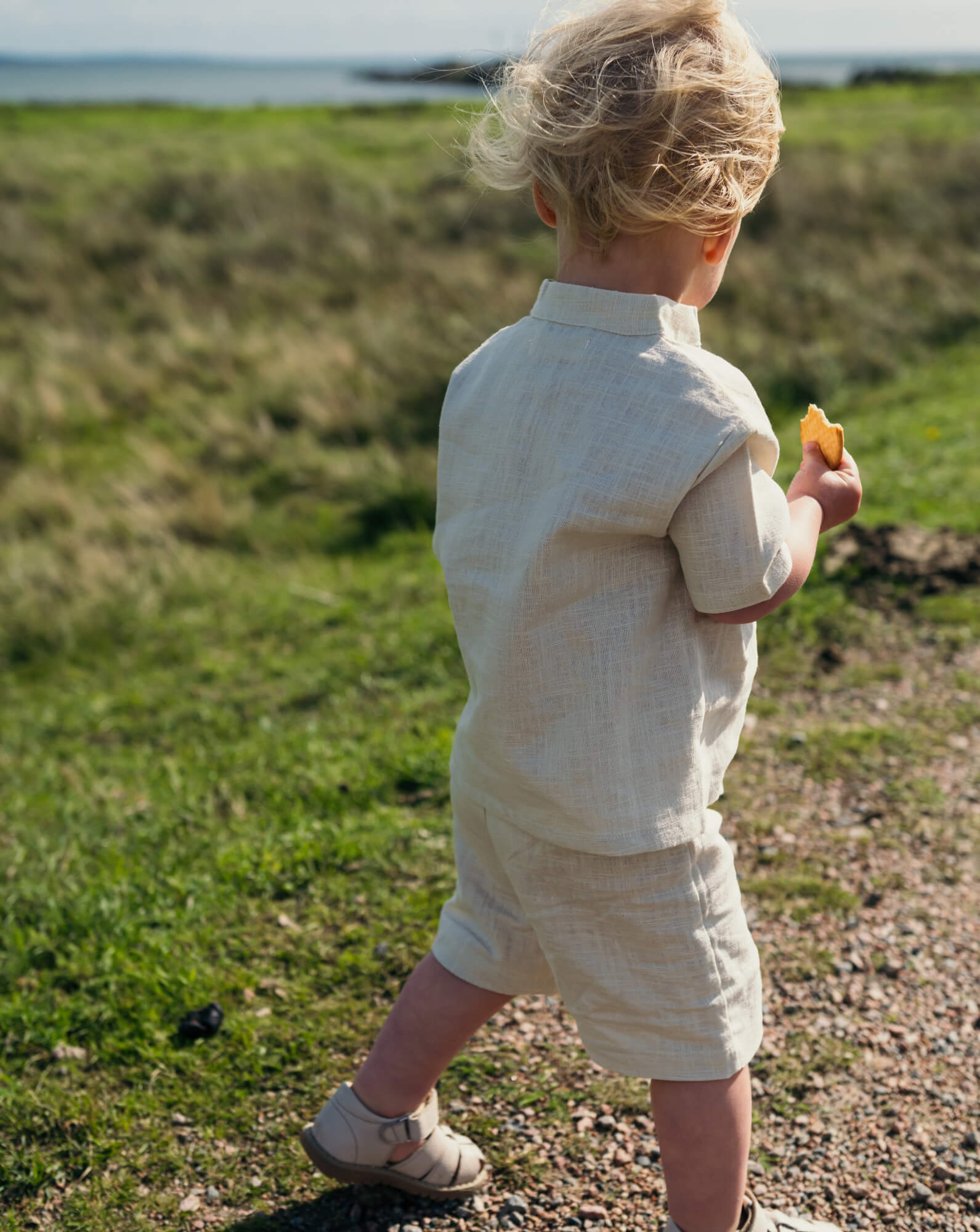 Back view of a baby wearing a beige short linen shirt and matching shorts while walking on a grassy path.