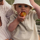 Baby wearing a beige short linen shirt while eating a fruit outdoors.