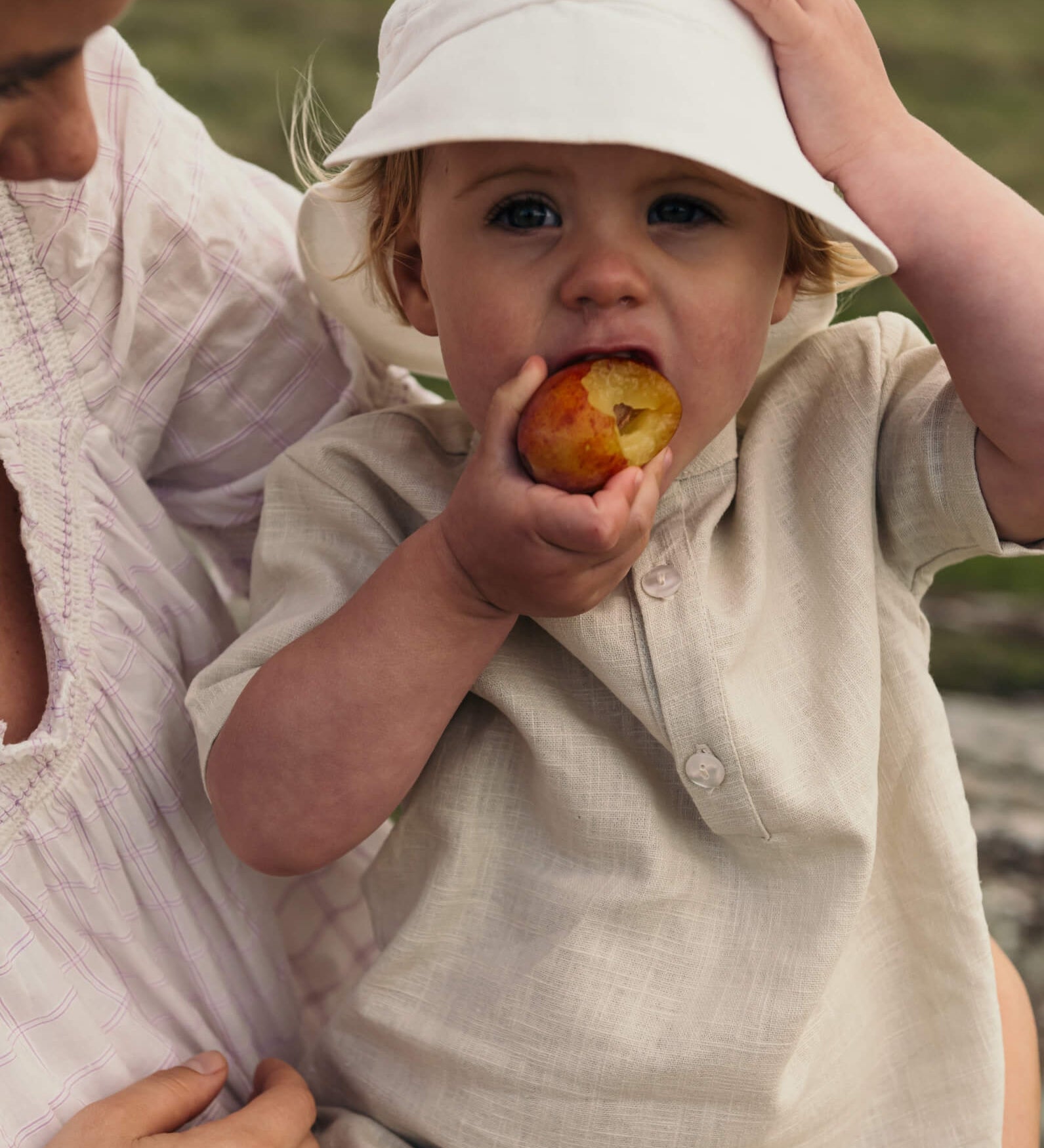 Baby wearing a beige short linen shirt while eating a fruit outdoors.