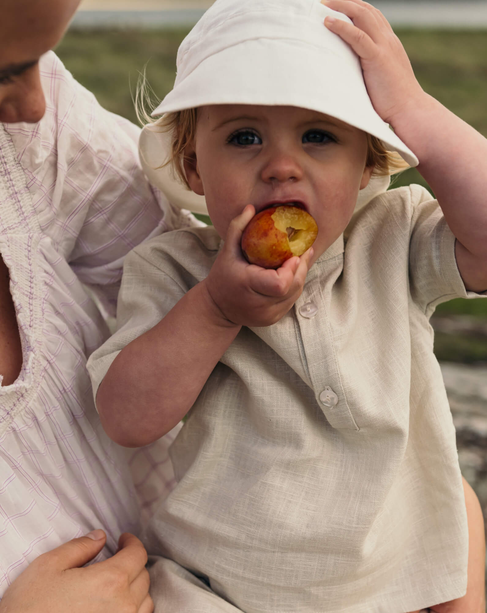 Baby wearing a beige short linen shirt while eating a fruit outdoors.