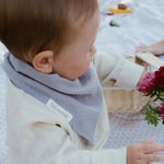 Soloveable organic grey muslin bib worn by a baby while reaching for flowers.