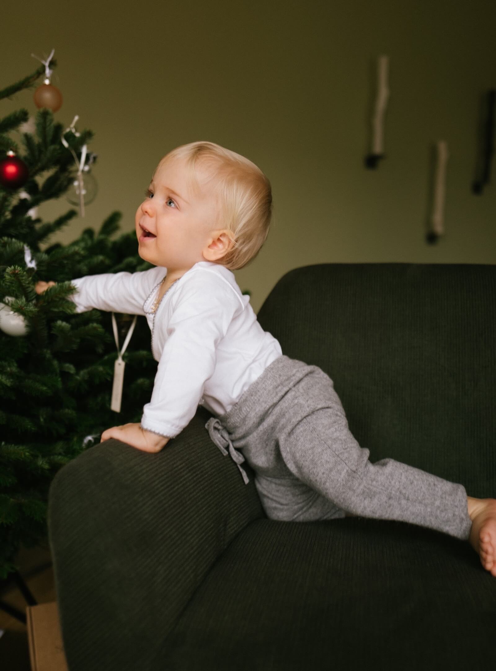 Baby in soft winter clothing reaching for the Christmas tree, wearing a white long-sleeve wrap bodysuit with crochet trim and grey cashmere pants.