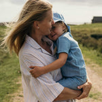 Mother holding toddler wearing a blue linen short-sleeve shirt and shorts outdoors.