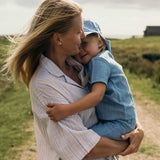 Mother holding toddler wearing a blue linen short-sleeve shirt and shorts outdoors.