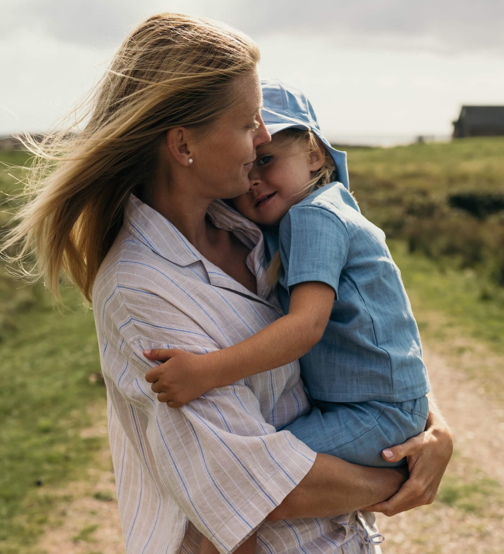 Mother holding toddler wearing a blue linen short-sleeve shirt and shorts outdoors.