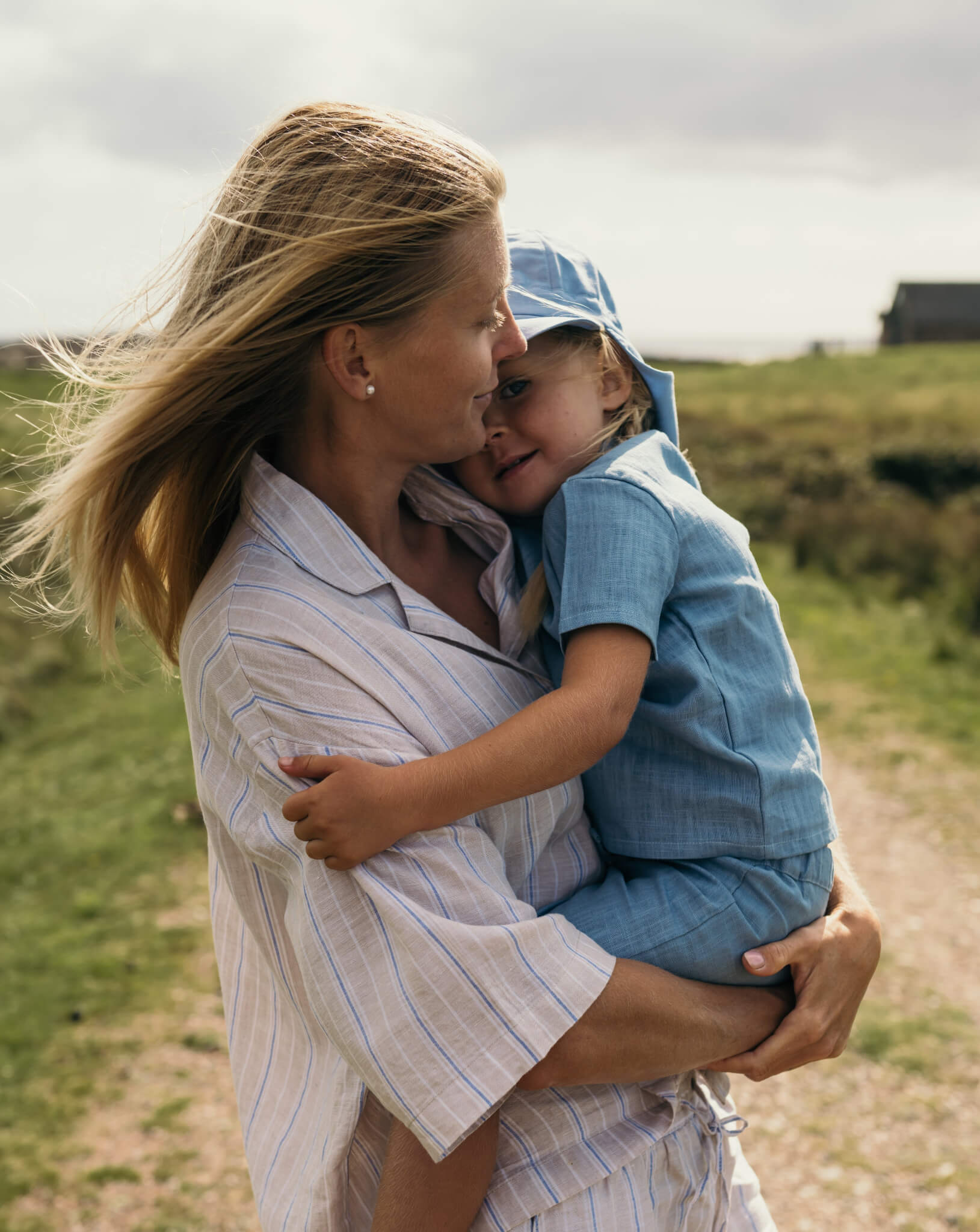Mother holding toddler wearing a blue linen short-sleeve shirt and shorts outdoors.