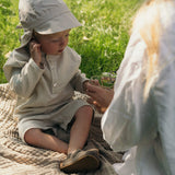 Toddler wearing a beige long-sleeve linen shirt and matching shorts while sitting on a blanket during a summer picnic.