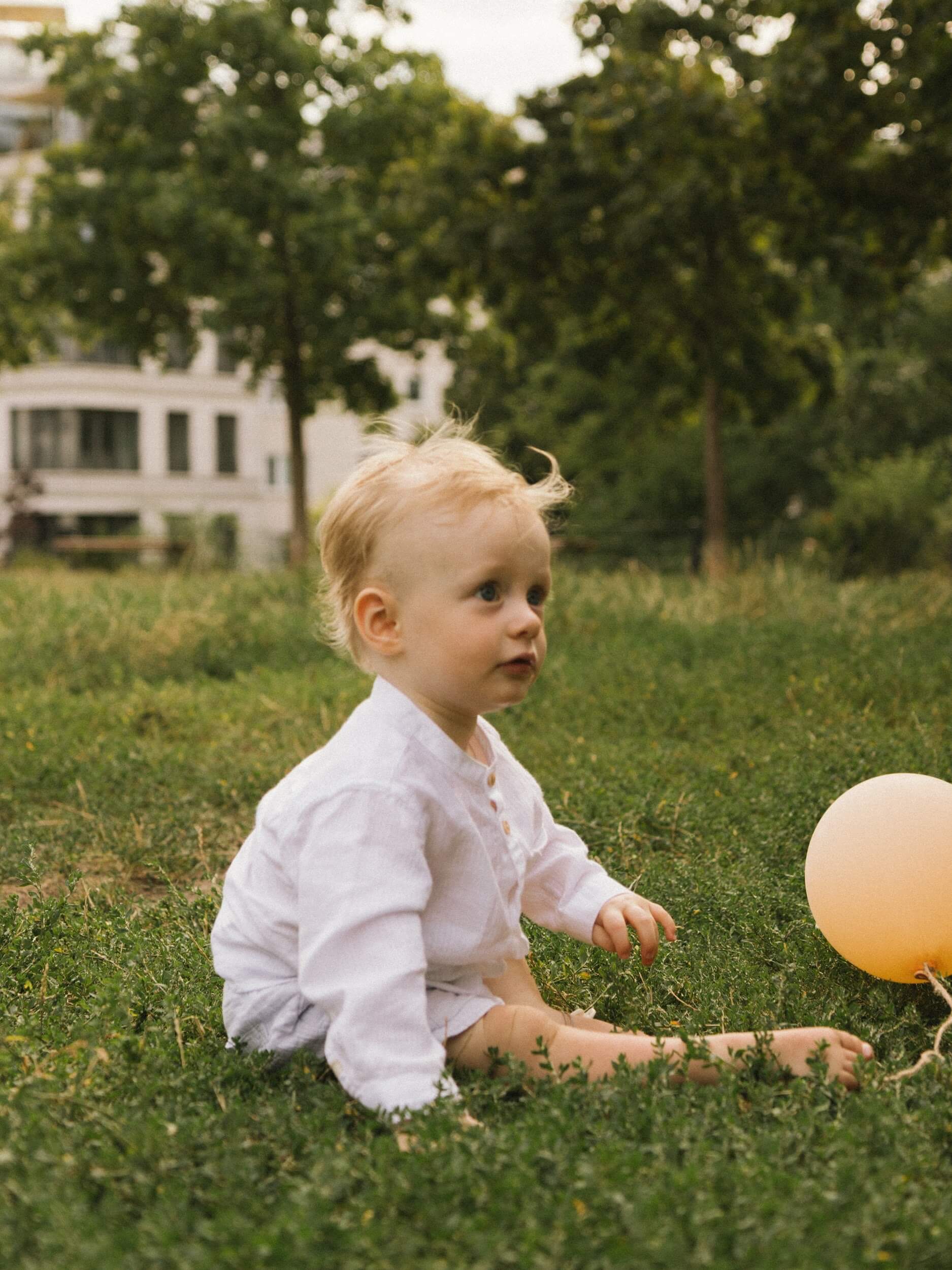 A toddler wearing a white organic linen shirt sitting on grass outdoors during playtime.