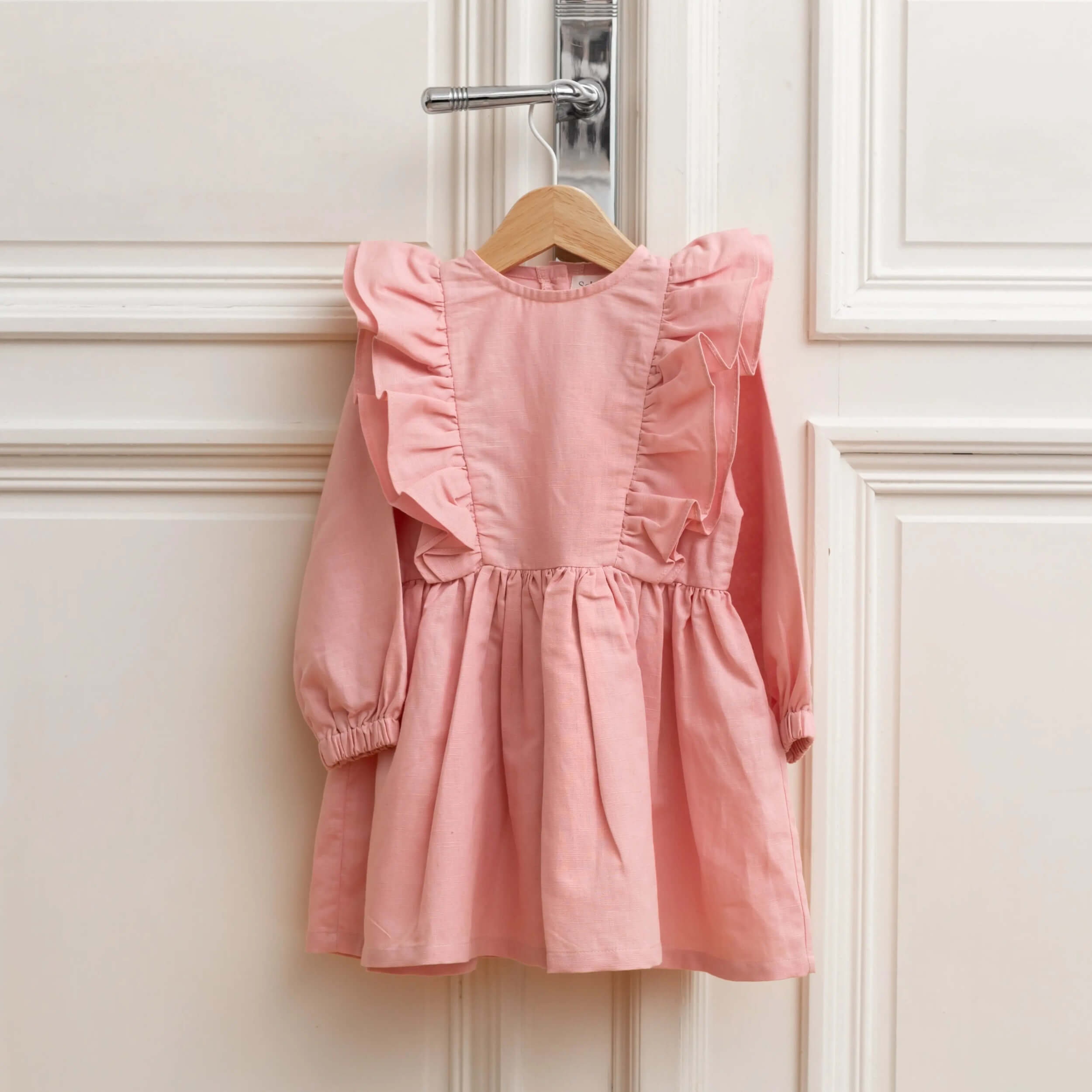 Pink linen girls dress with long sleeves and ruffle details hanging on a wooden hanger