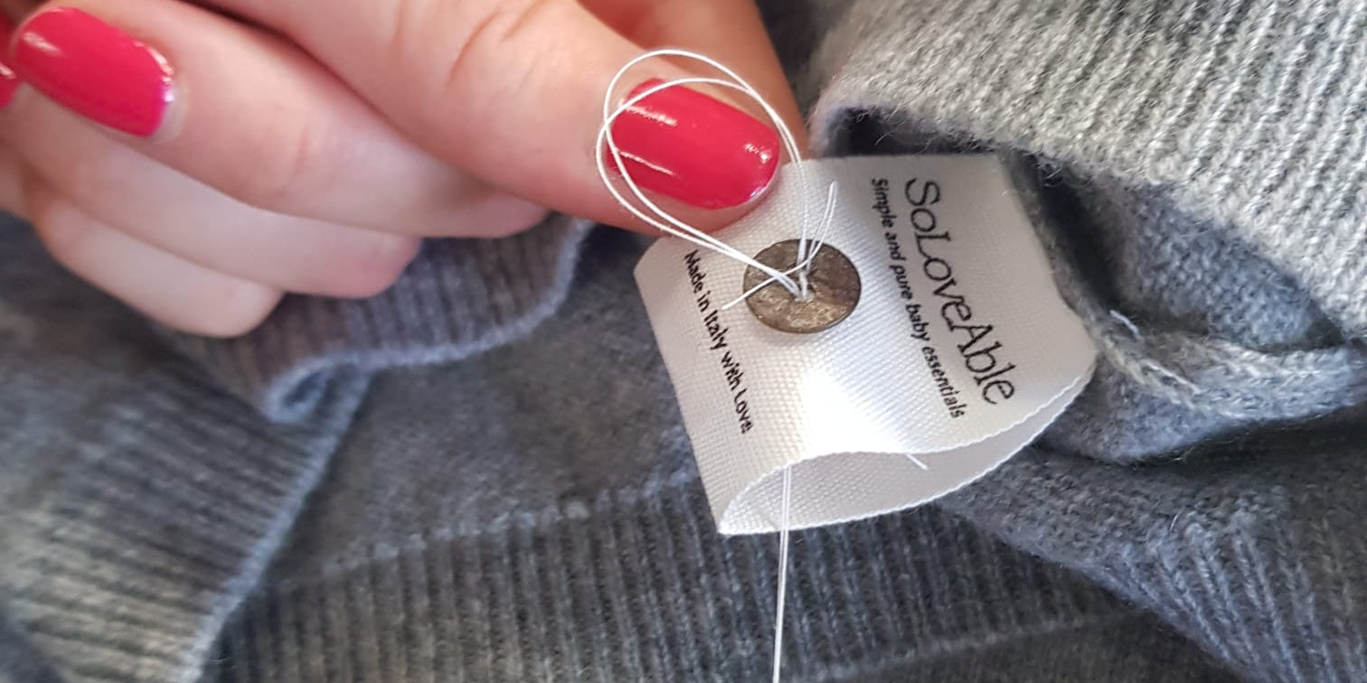 Close-up of a hand sewing a button and label onto a grey cashmere garment, representing handcrafted quality and careful finishing by SoLoveAble.