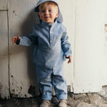 Toddler wearing a blue linen shirt and matching linen pants, standing outdoors against a rustic white wooden wall.