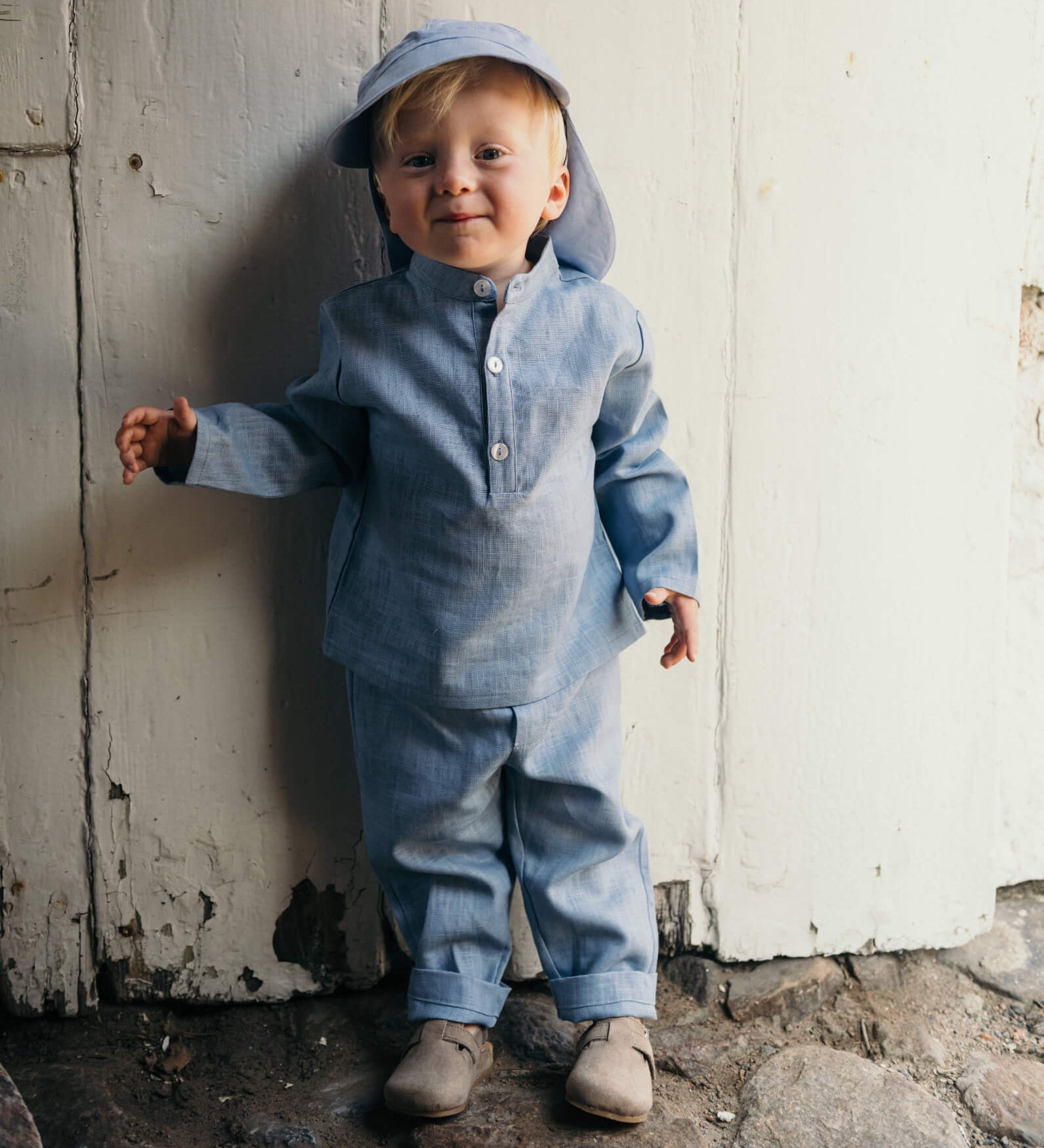 Toddler wearing a blue linen shirt and matching linen pants, standing outdoors against a rustic white wooden wall.