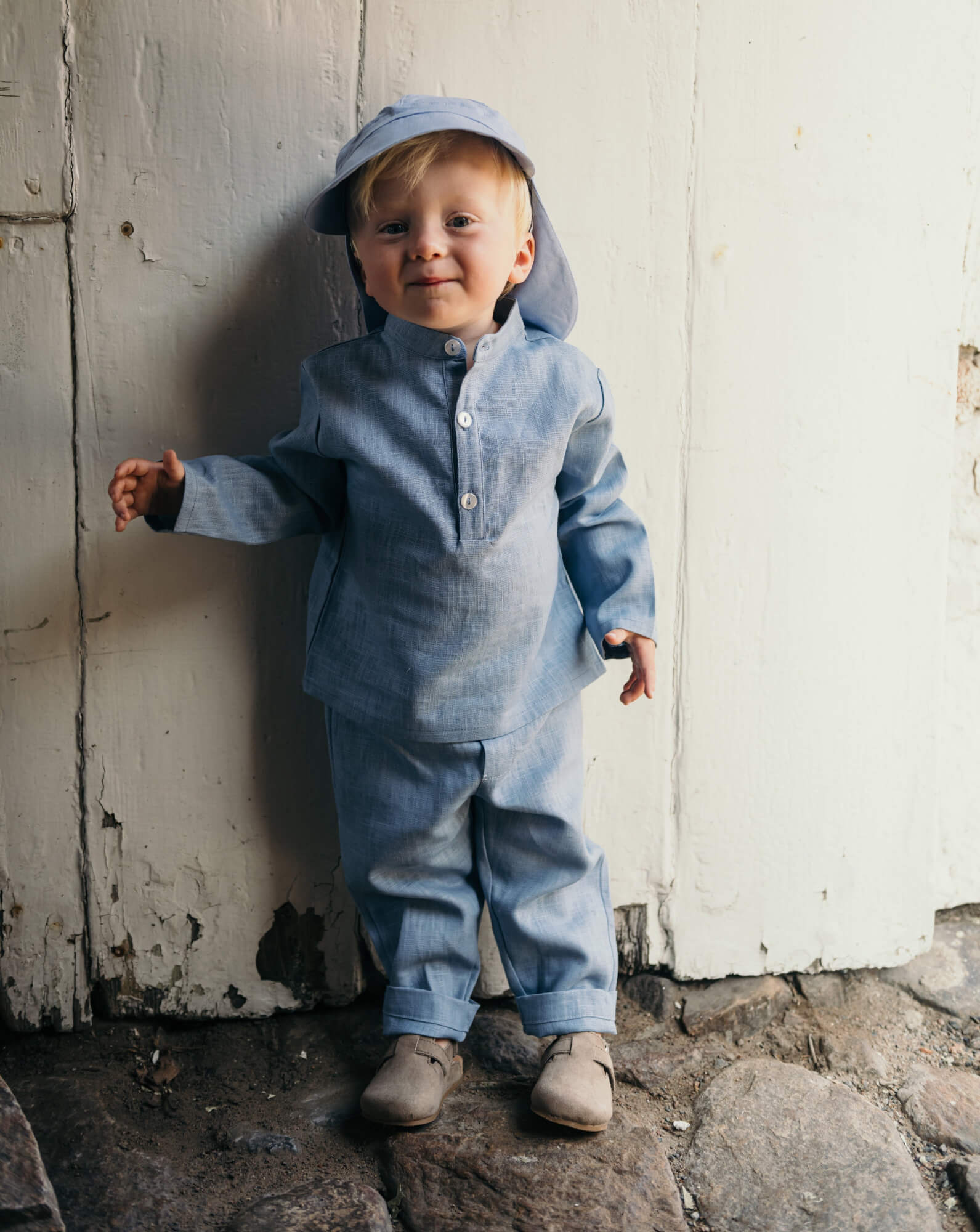Toddler wearing a blue linen shirt and matching linen pants, standing outdoors against a rustic white wooden wall.