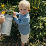Toddler wearing a blue linen shirt and beige linen shorts, holding a watering can in a garden.