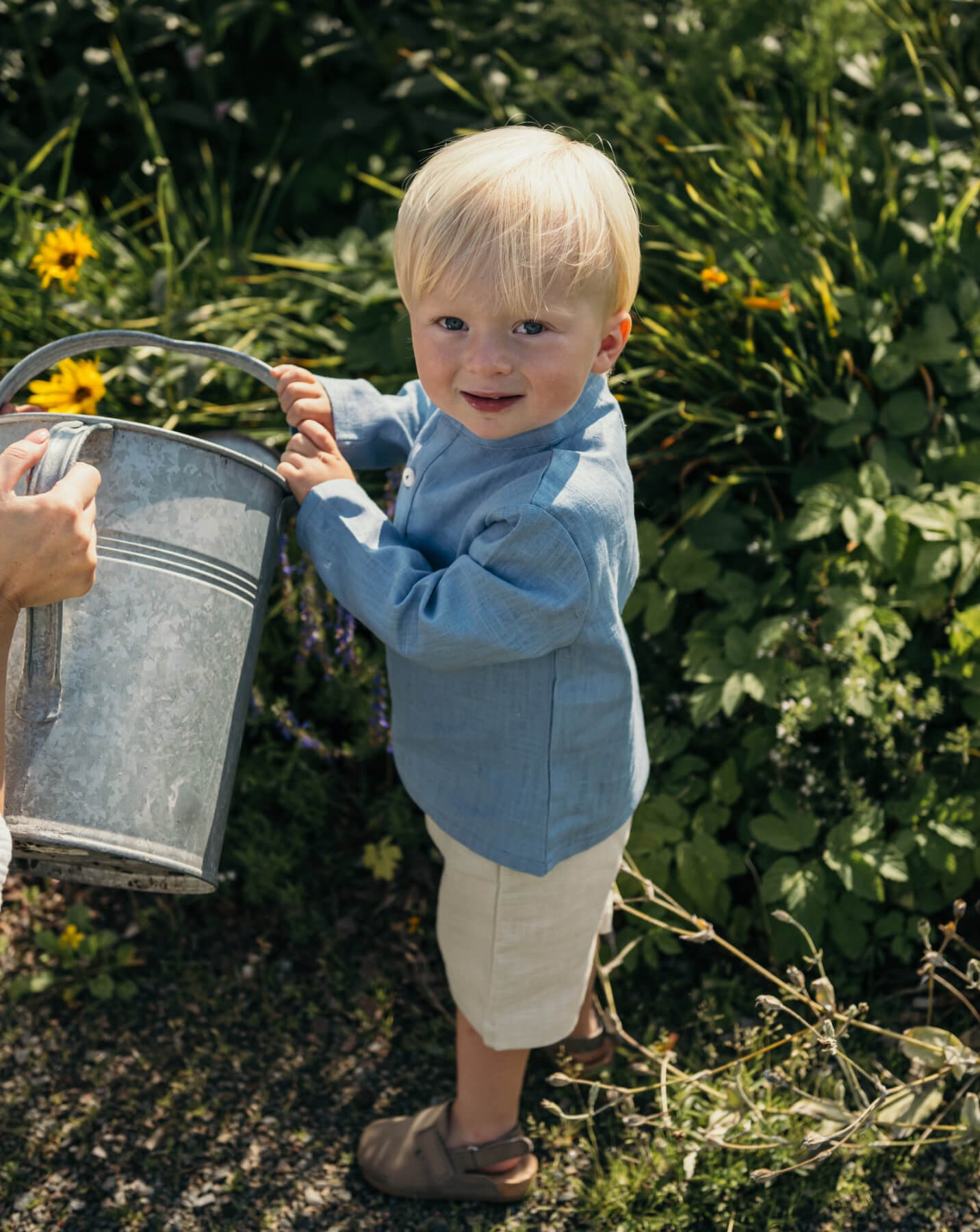 Toddler wearing a blue linen shirt and beige linen shorts, holding a watering can in a garden.