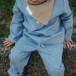 Toddler wearing a light blue linen shirt and pants with a beige muslin bib, sitting outdoors on a stone wall.
