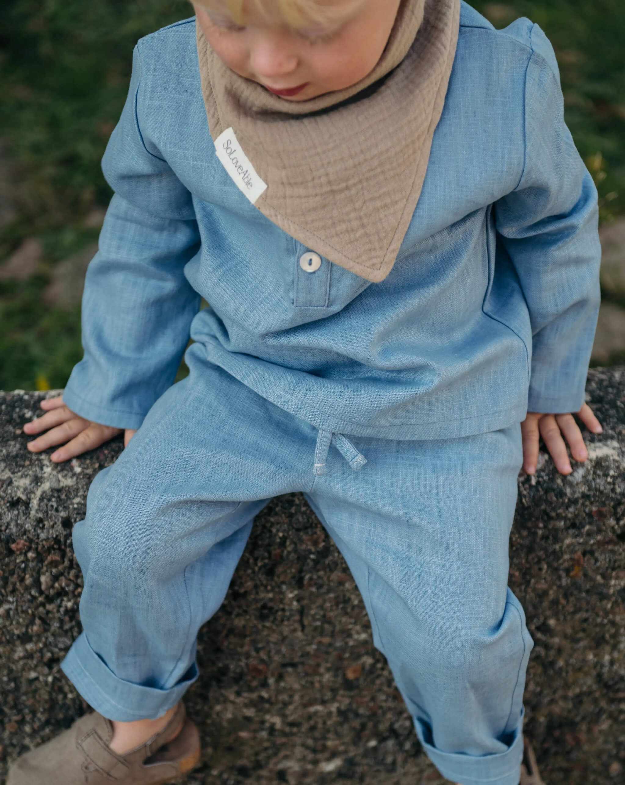Toddler wearing a light blue linen shirt and pants with a beige muslin bib, sitting outdoors on a stone wall.
