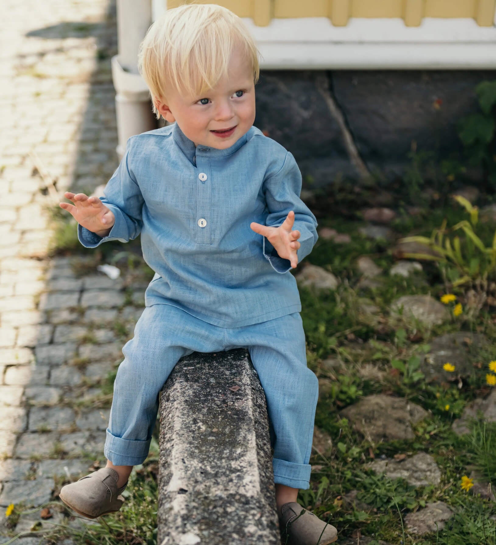 Toddler wearing a light blue linen shirt and matching linen pants sitting outdoors on a stone wall.