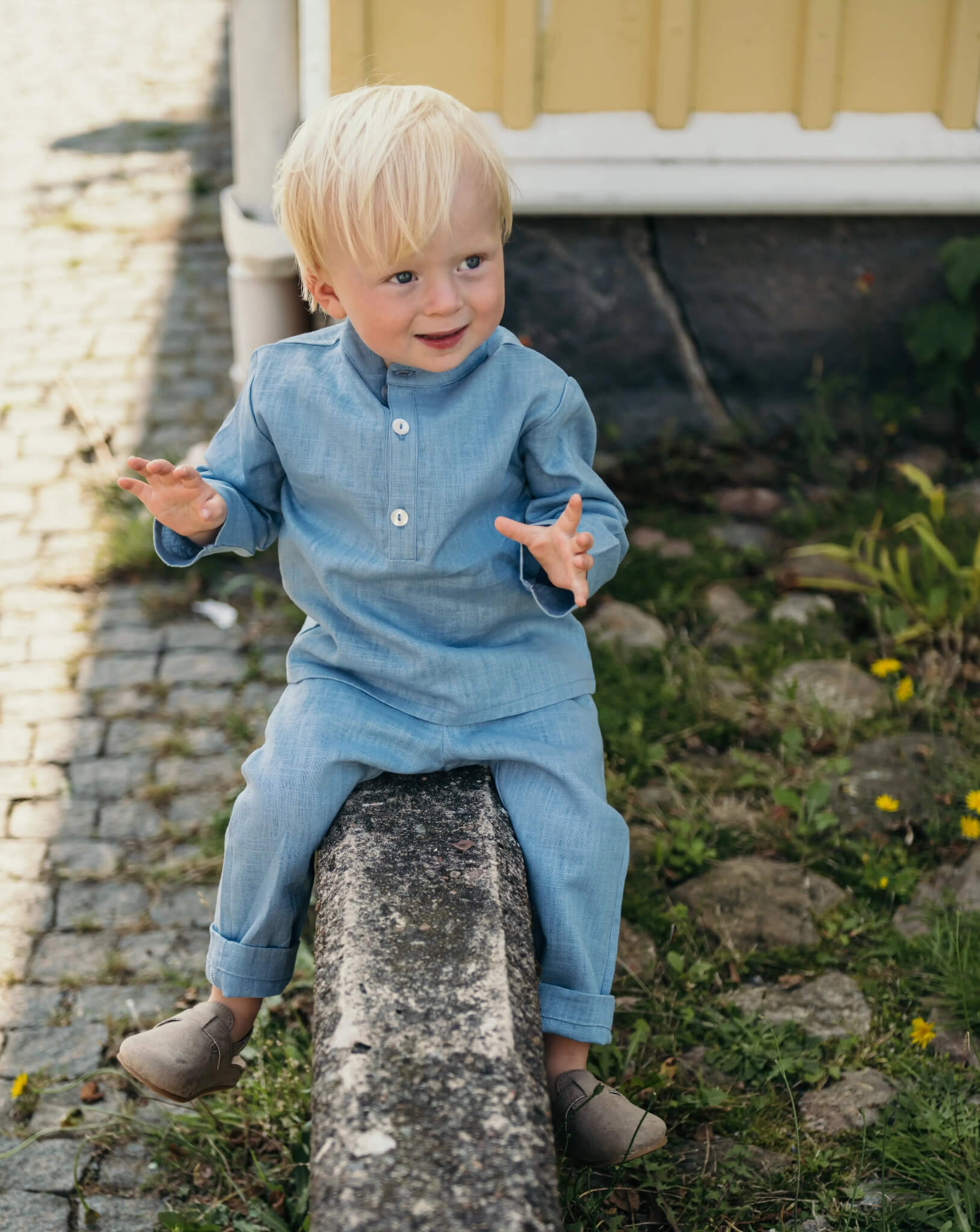 Toddler wearing a light blue linen shirt and matching linen pants sitting outdoors on a stone wall.