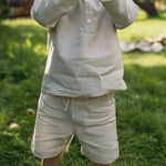 Toddler wearing short beige linen pants and a matching linen shirt outdoors on a sunny day.