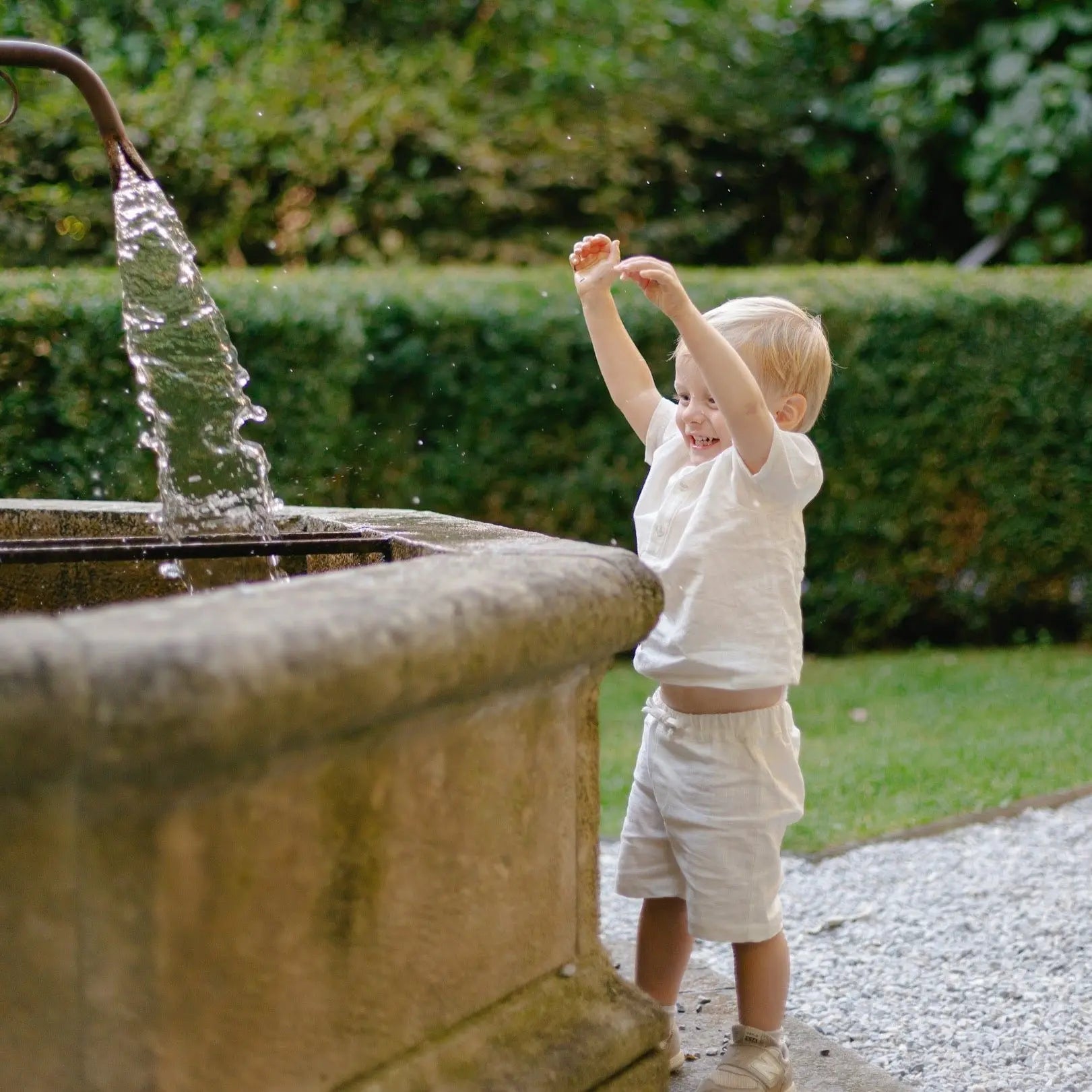 Toddler playing outdoors wearing short white linen pants and a matching shirt near a fountain.