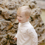 Toddler wearing a long-sleeve white linen shirt outdoors, smiling near a stone fountain.
