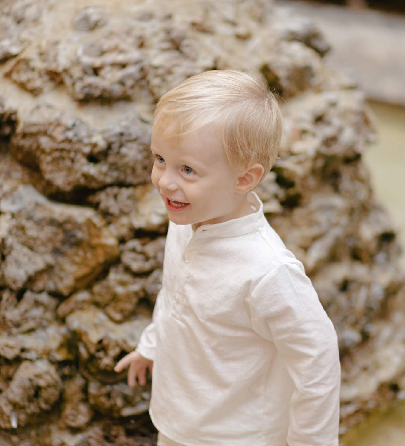 Toddler wearing a long-sleeve white linen shirt outdoors, smiling near a stone fountain.
