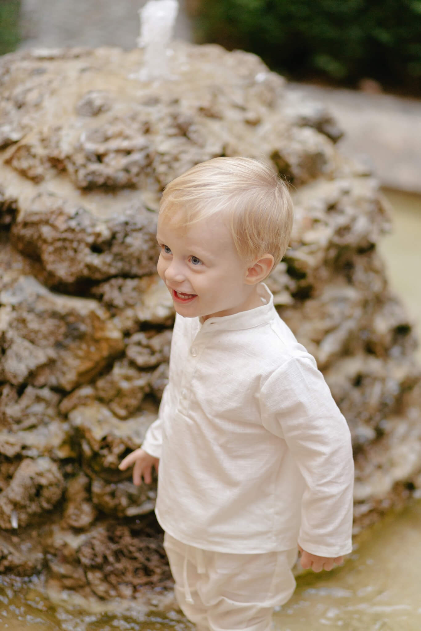 Toddler wearing a long-sleeve white linen shirt outdoors, smiling near a stone fountain.