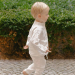 Toddler in white linen pants and matching shirt, walking away outdoors.