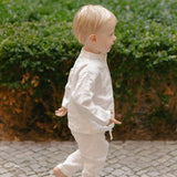 Toddler in white linen pants and matching shirt, walking away outdoors.