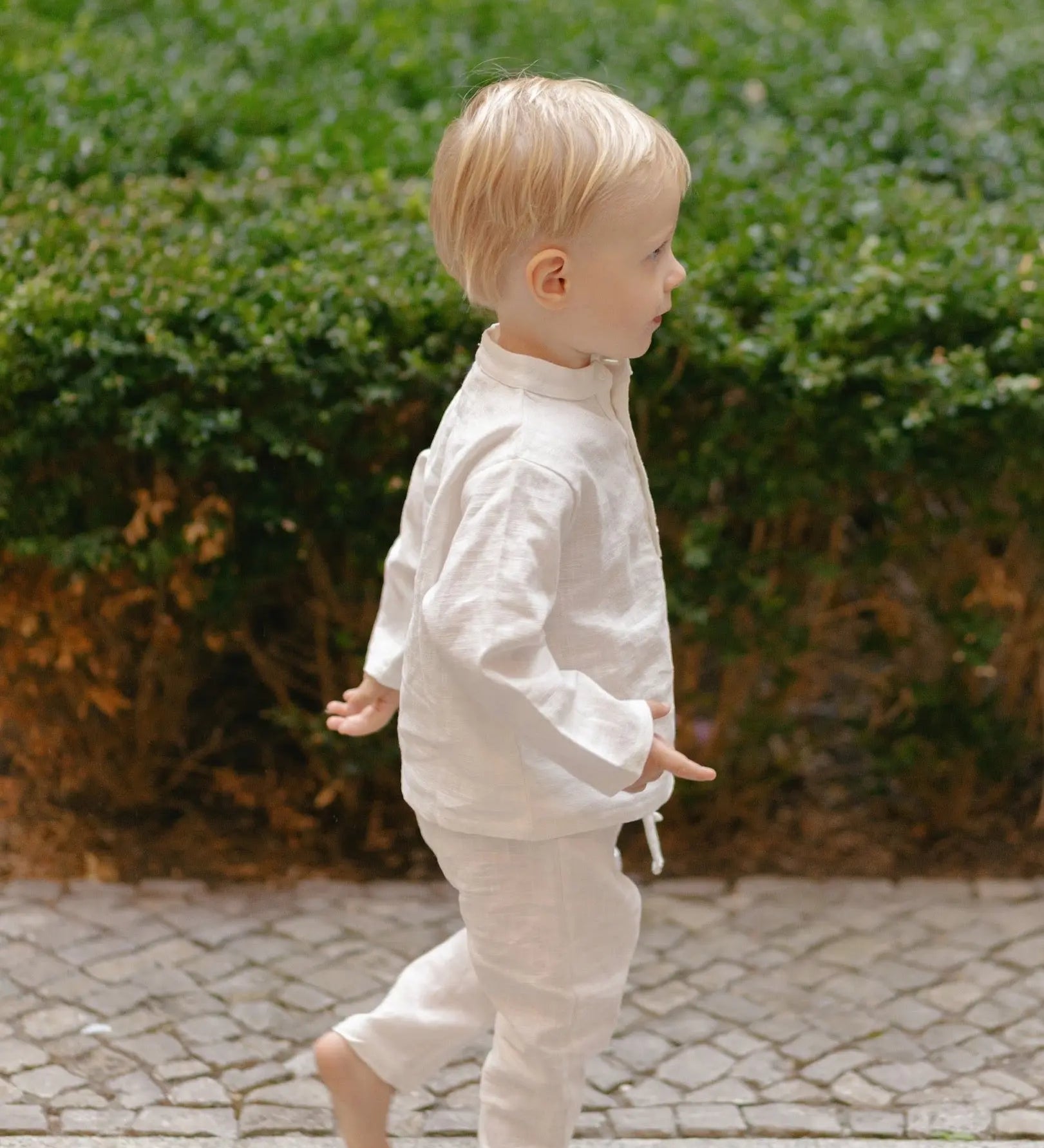 Toddler in white linen pants and matching shirt, walking away outdoors.