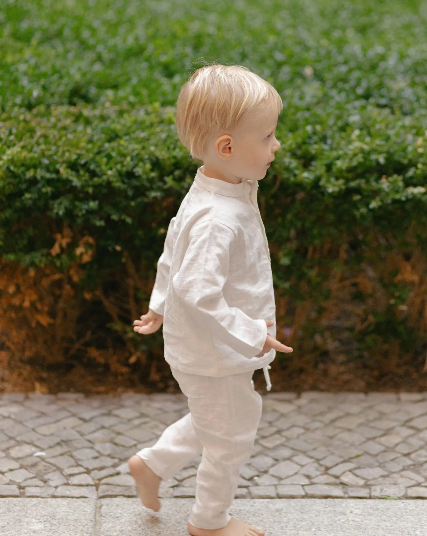 Toddler in white linen pants and matching shirt, walking away outdoors.