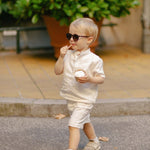 Toddler walking outdoors wearing a white linen shirt and matching shorts while eating ice cream.