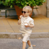 Toddler walking outdoors wearing a white linen shirt and matching shorts while eating ice cream.