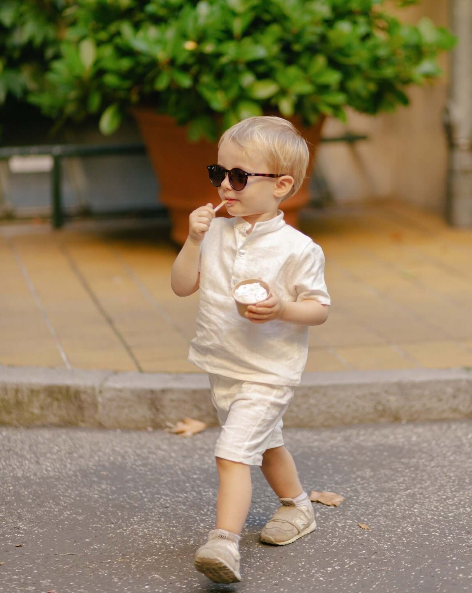 Toddler walking outdoors wearing a white linen shirt and matching shorts while eating ice cream.