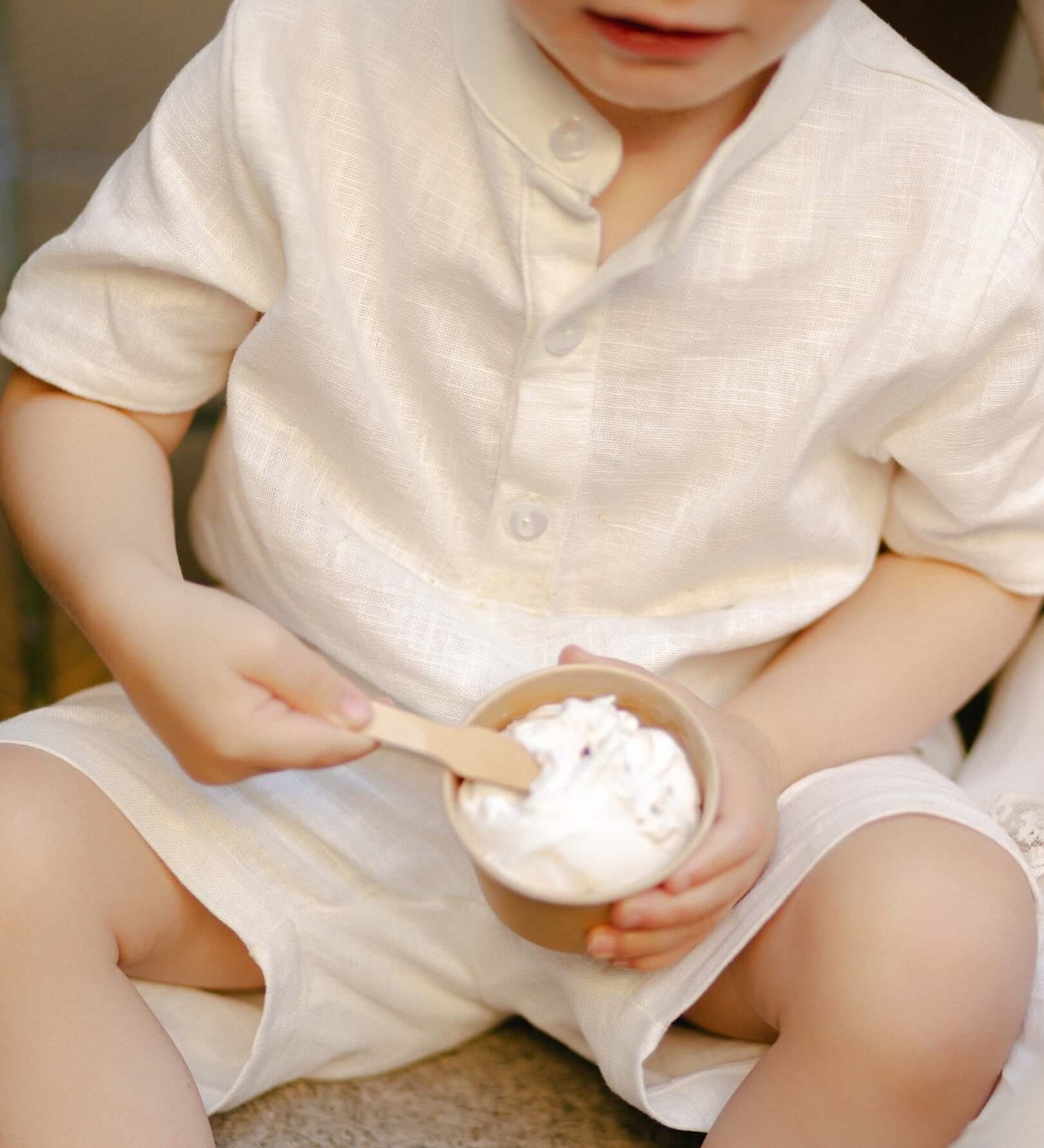Toddler wearing a white linen short-sleeve shirt eating ice cream outdoors.