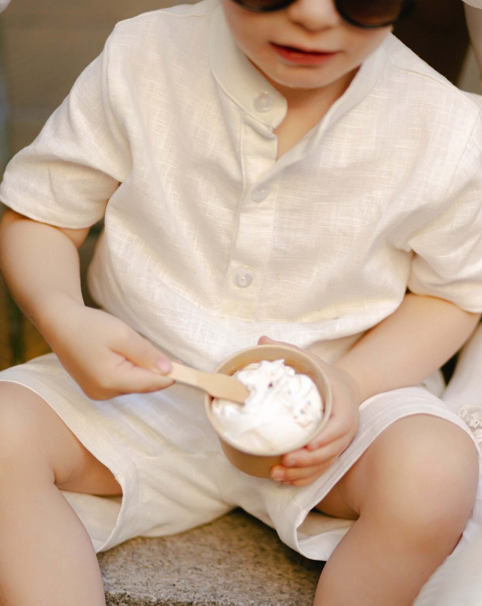 Toddler wearing a white linen short-sleeve shirt eating ice cream outdoors.