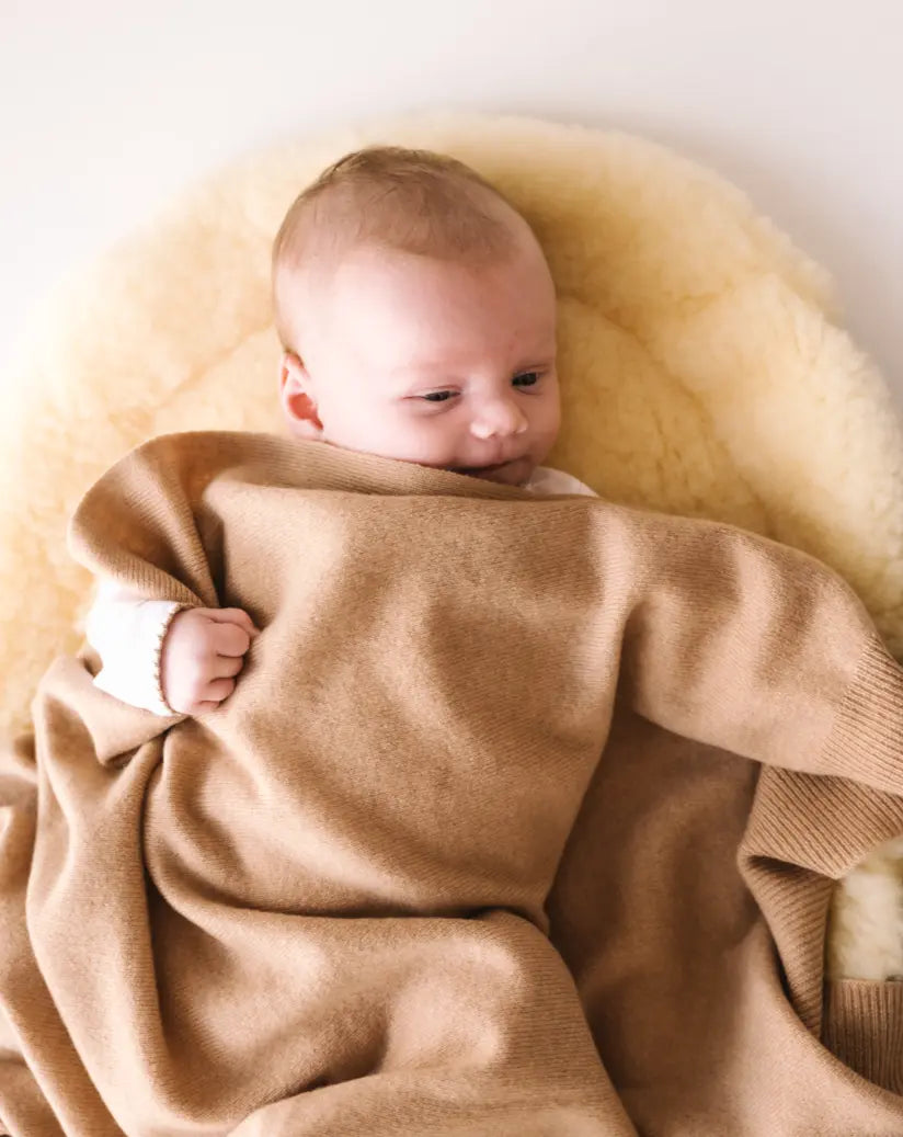 Baby cuddled in a caramel brown cashmere blanket, lying on a soft cream pad.
