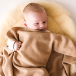 Baby cuddled in a caramel brown cashmere blanket, lying on a soft cream pad.