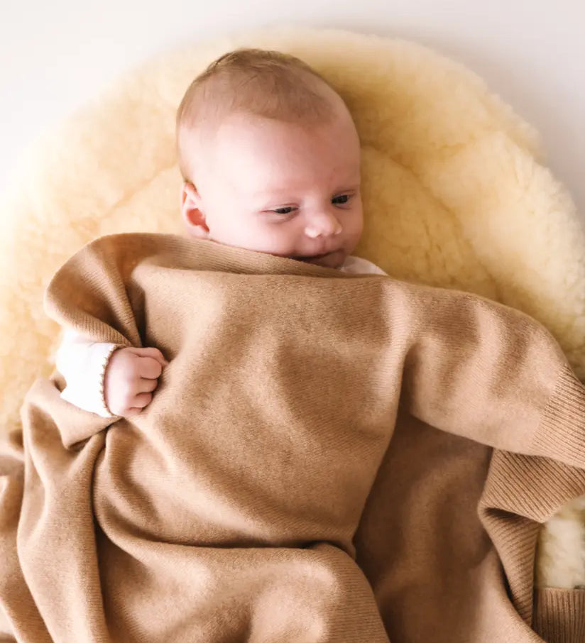 Baby cuddled in a caramel brown cashmere blanket, lying on a soft cream pad.